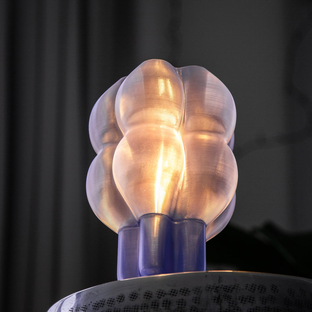 A modern, glowing translucent bulb-shaped table lamp sits on a hammered silver metal stool, surrounded by large green houseplant leaves in a dimly lit interior setting.