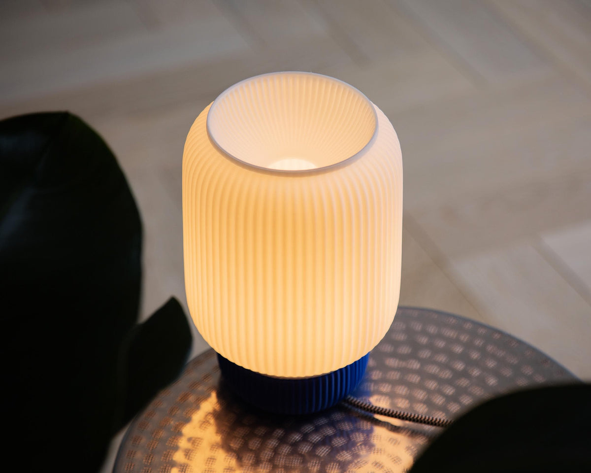 A modern three-tiered ribbed table lamp glowing with a warm cream color, set on a dark surface with a blurred background.