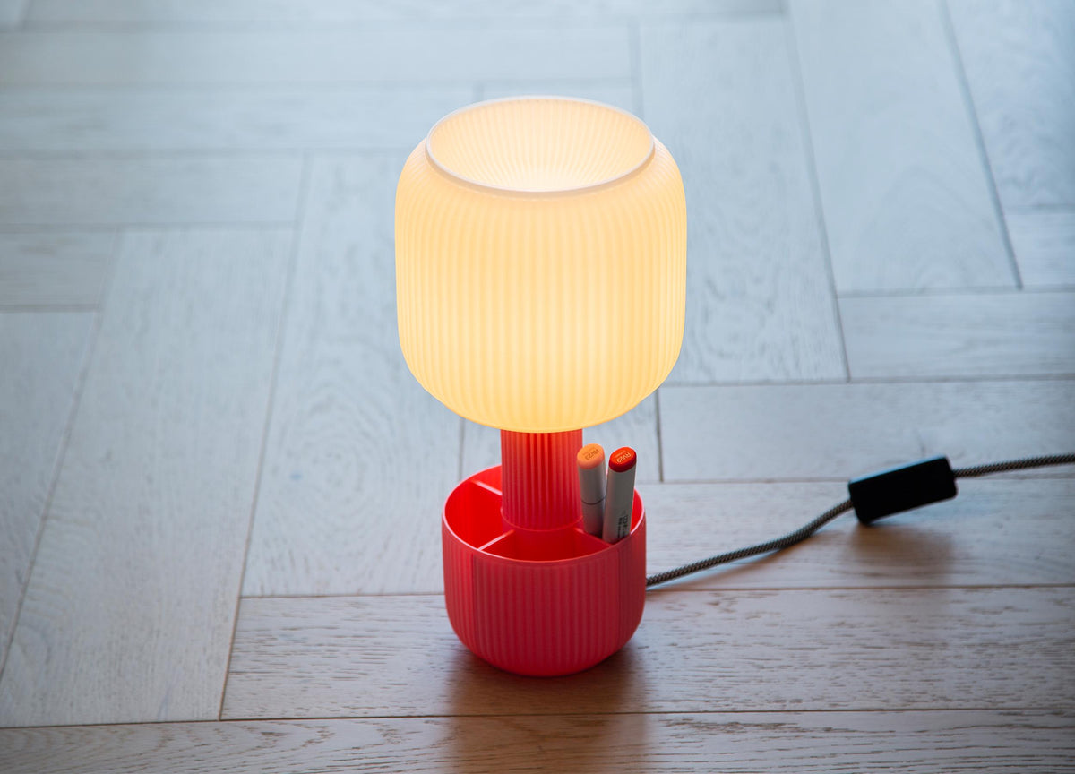 A ribbed translucent white plastic lampshade with a red fluted base, displayed on a wood surface against a neutral gray background.