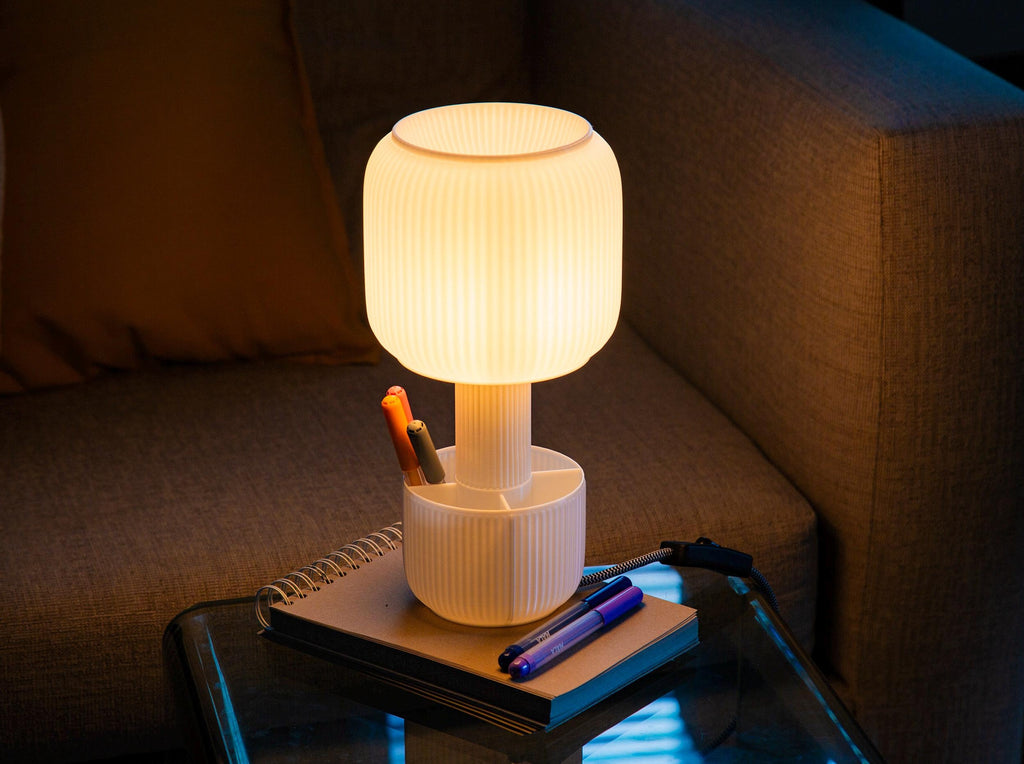 A modern light with a flared, ruffled shade made of warm white or cream-colored material, glowing with a soft yellow light against a dark gray background.