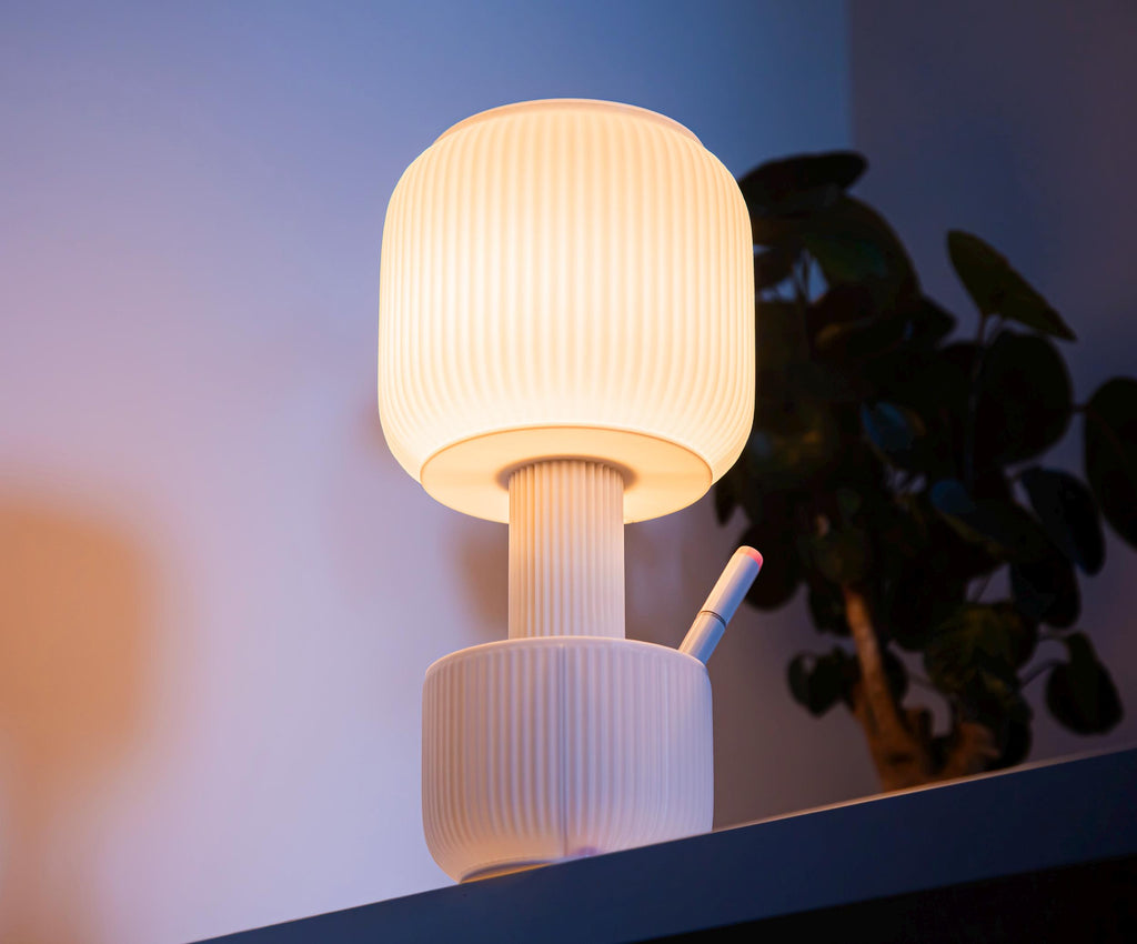A modern light with a flared, pleated shade glowing with a warm yellow light, suspended above a round table next to a notebook.