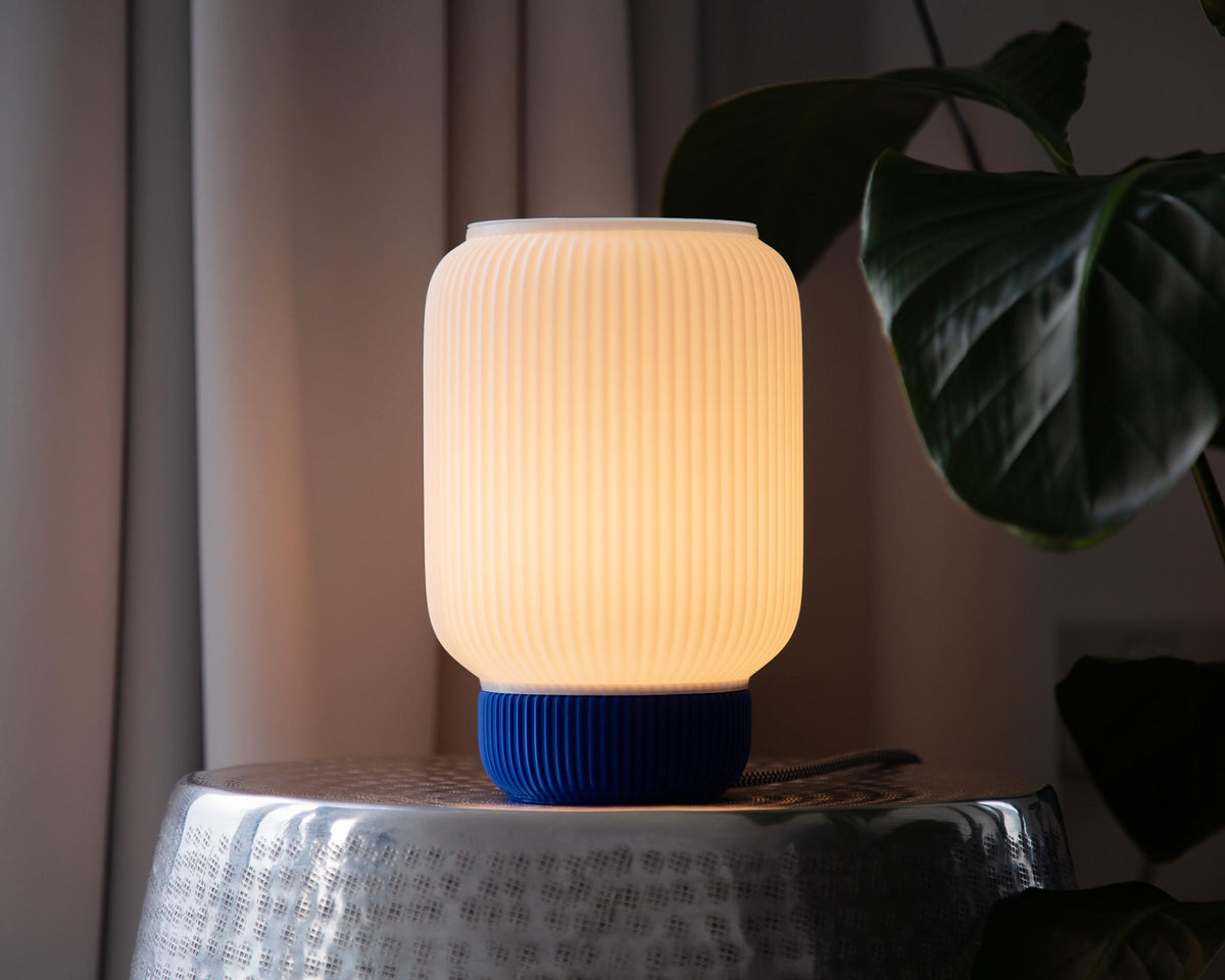 A ribbed cylindrical table lamp with a glowing warm white shade and a dark blue ribbed base, styled on a metallic side table with a plant leaf in the background.