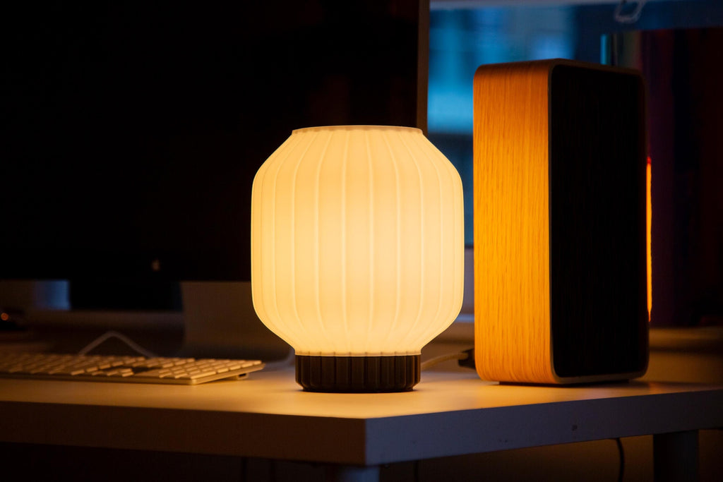 A glowing cylindrical table lamp with a pleated white shade and dark base sits on a desk next to a tall wooden speaker in a dim room.