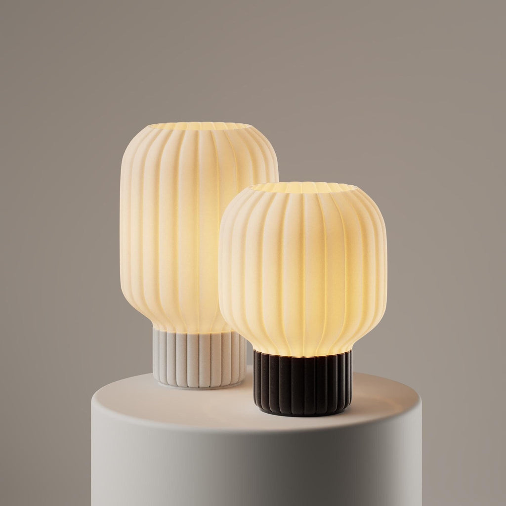A modern pendant light with a flared, ruffled shade made of warm white or cream-colored material, glowing with a soft yellow light against a dark gray background.