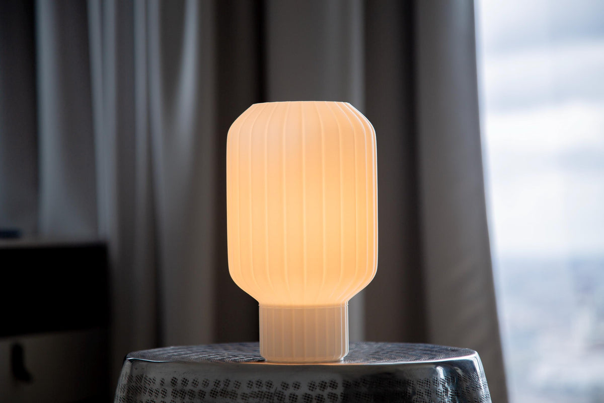A modern, glowing translucent bulb-shaped table lamp sits on a hammered silver metal stool, surrounded by large green houseplant leaves in a dimly lit interior setting.