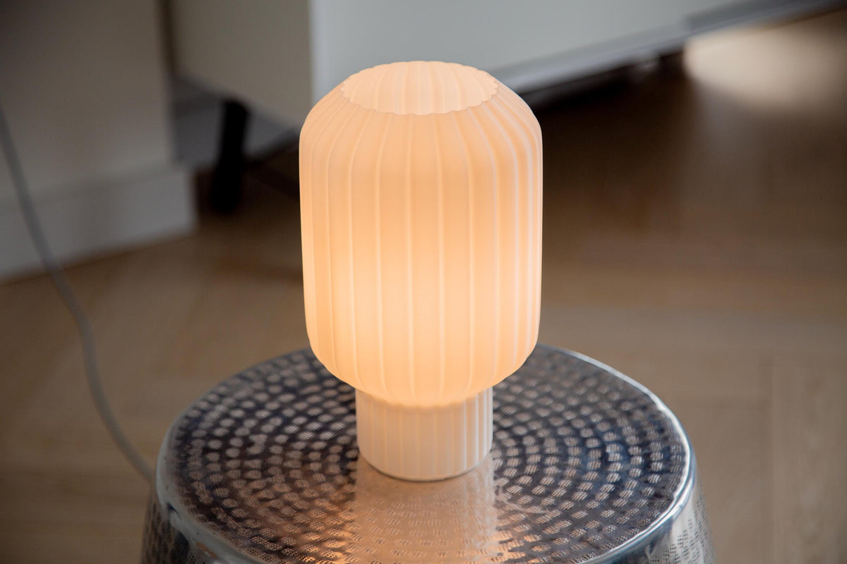 A ribbed, cylindrical table lamp glowing with a warm cream-white light, resting on a hammered metal stool in a softly blurred indoor setting.