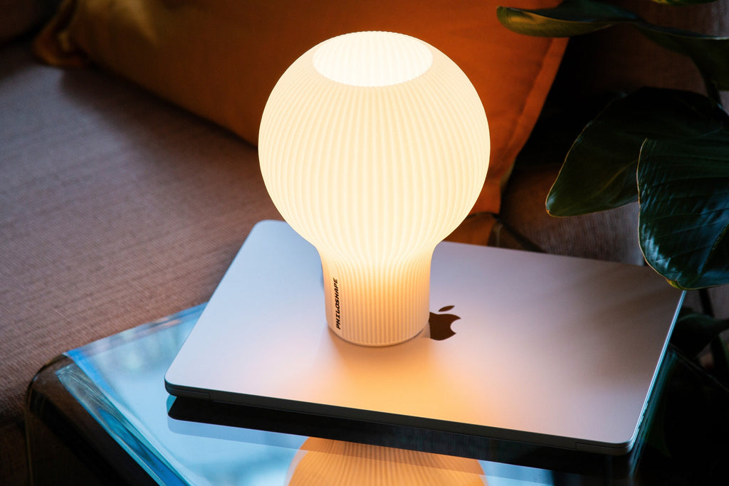 A modern pendant light with a flared, pleated shade glowing with a warm yellow light, suspended above a round table next to a notebook.