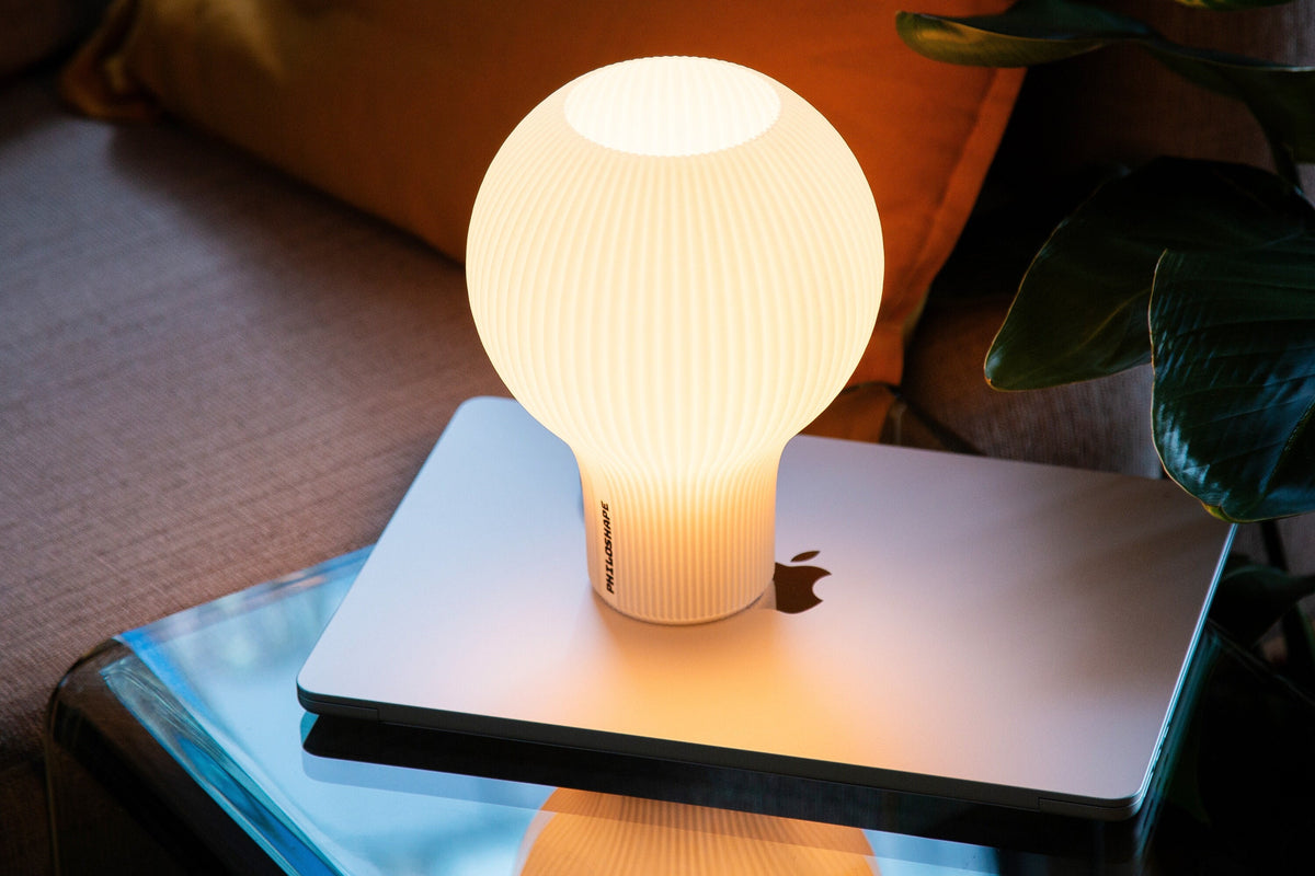 A modern pendant light with a flared, pleated shade glowing with a warm yellow light, suspended above a round table next to a notebook.