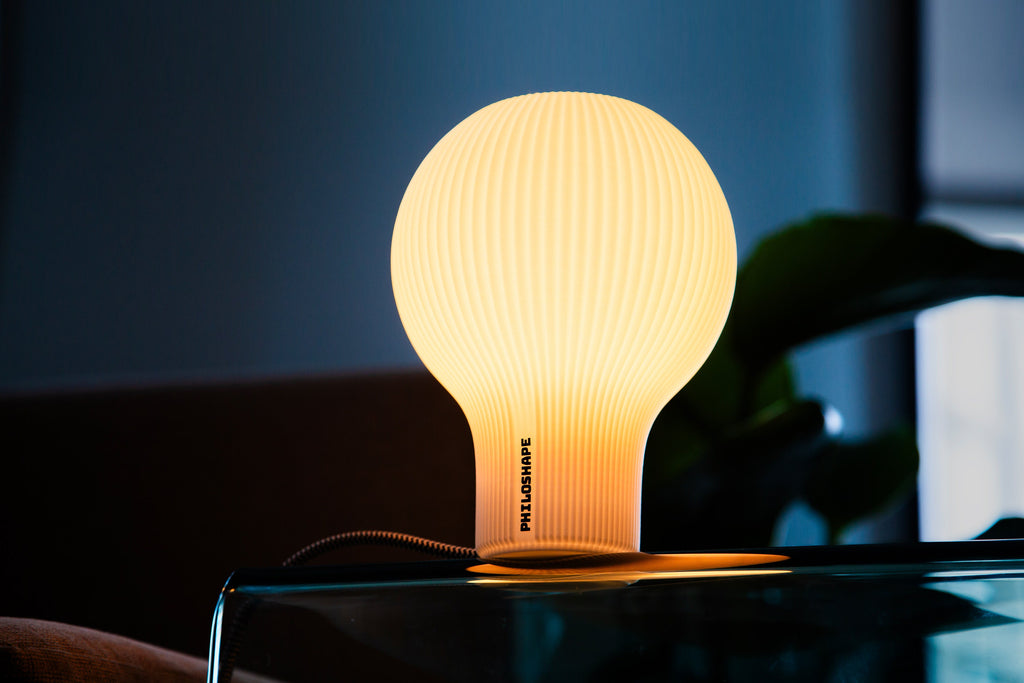 A modern table light with a flared, ruffled shade made of warm white or cream-colored material, glowing with a soft yellow light against a dark gray background.