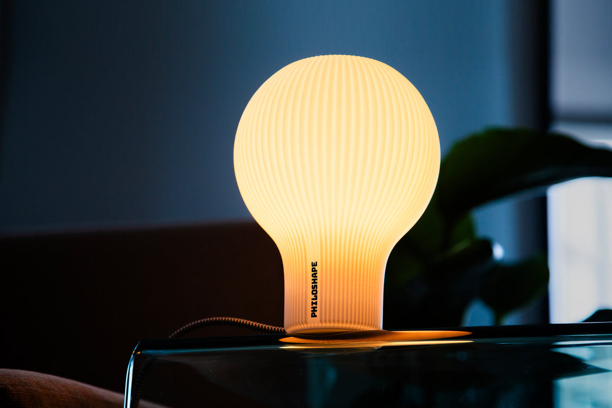 A modern table light with a flared, ruffled shade made of warm white or cream-colored material, glowing with a soft yellow light against a dark gray background.