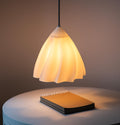 A modern pendant light with a flared, pleated shade glowing with a warm yellow light, suspended above a round table next to a notebook.