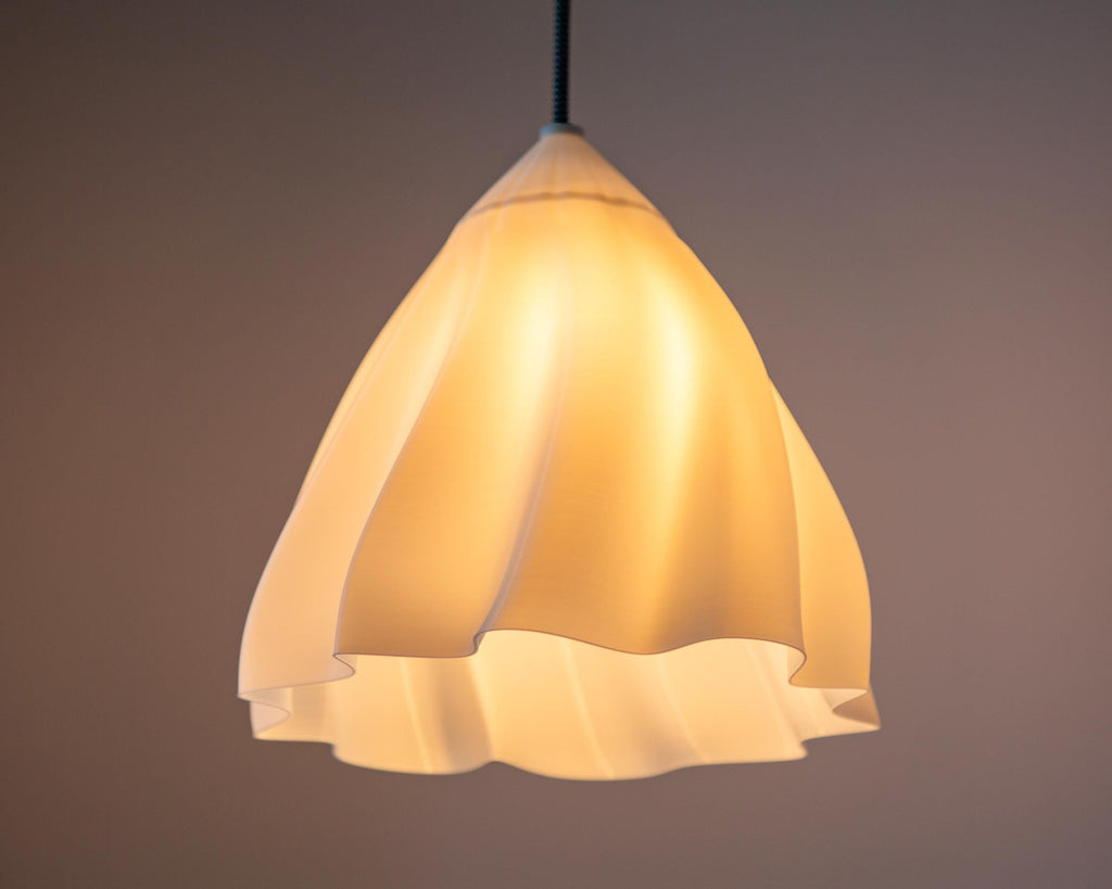 A modern pendant light with a flared, ruffled shade made of warm white or cream-colored material, glowing with a soft yellow light against a dark brown background.