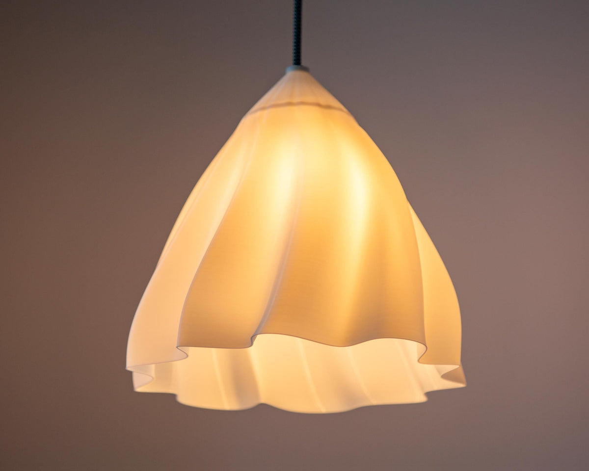 A modern pendant light with a flared, ruffled shade made of warm white or cream-colored material, glowing with a soft yellow light against a dark brown background.