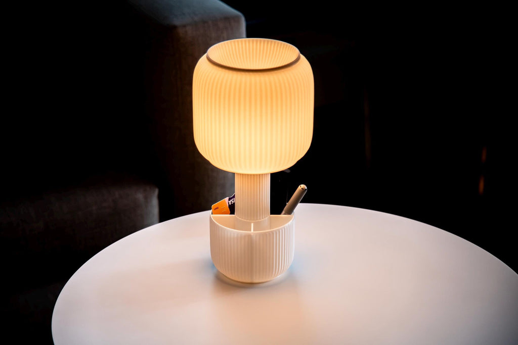 A modern ribbed palstic table lamp with a glowing warm yellow shade and a white pen holder base, sitting on a white round table in a dark interior setting.