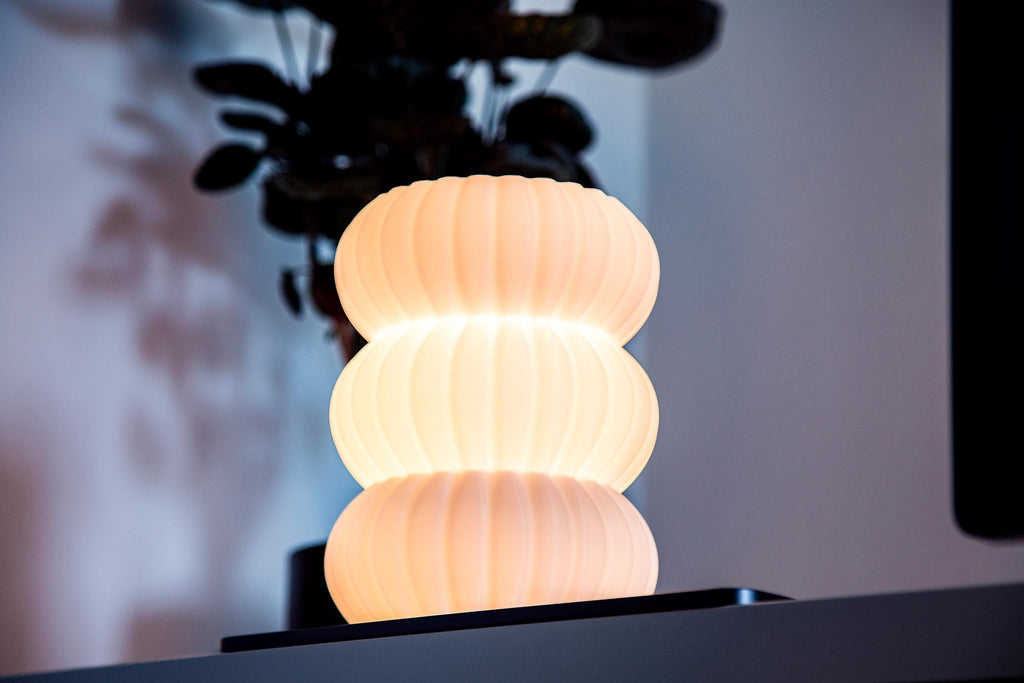 A modern, glowing translucent bulb-shaped table lamp sits on a hammered silver metal stool, surrounded by large green houseplant leaves in a dimly lit interior setting.