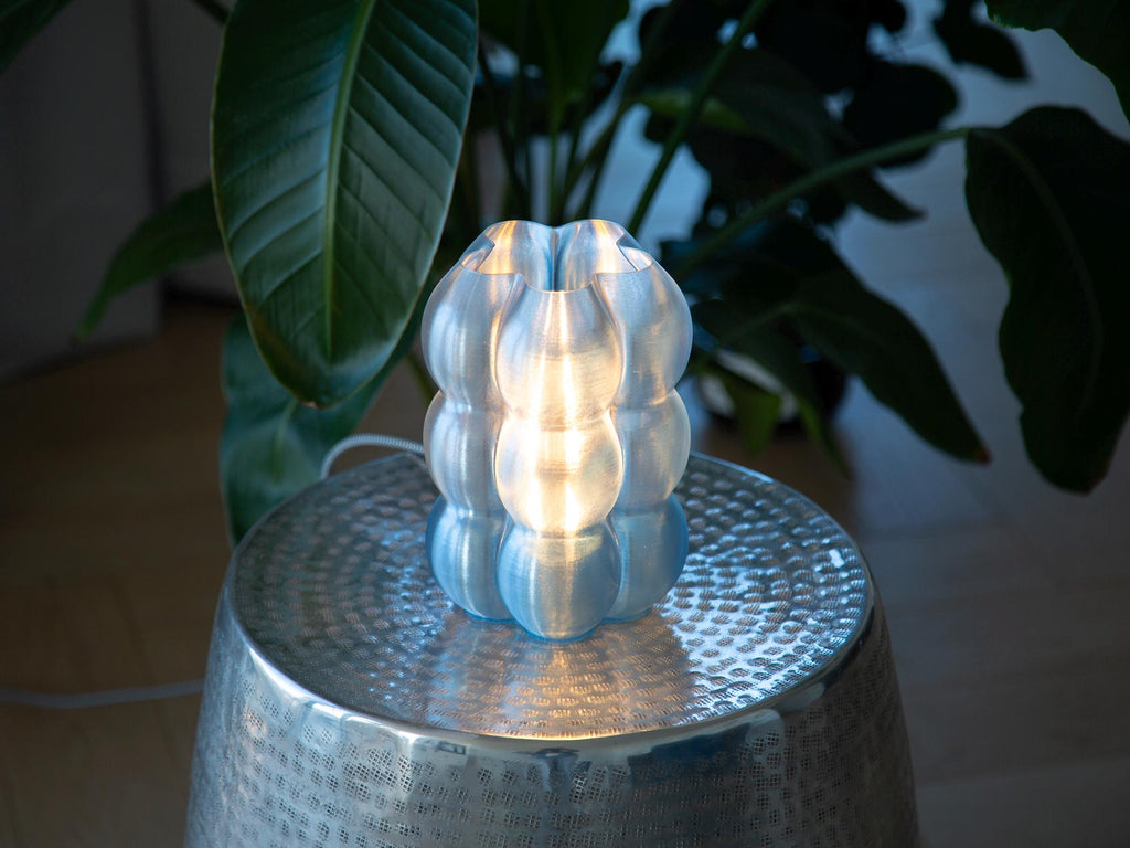A modern, glowing translucent bulb-shaped table lamp sits on a hammered silver metal stool, surrounded by large green houseplant leaves in a dimly lit interior setting.