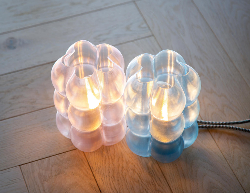 Two modern bubble cluster table lamps on a light wood floor, one glowing with warm pinkish light and the other with cool blue light, with a metal cord visible on the right.