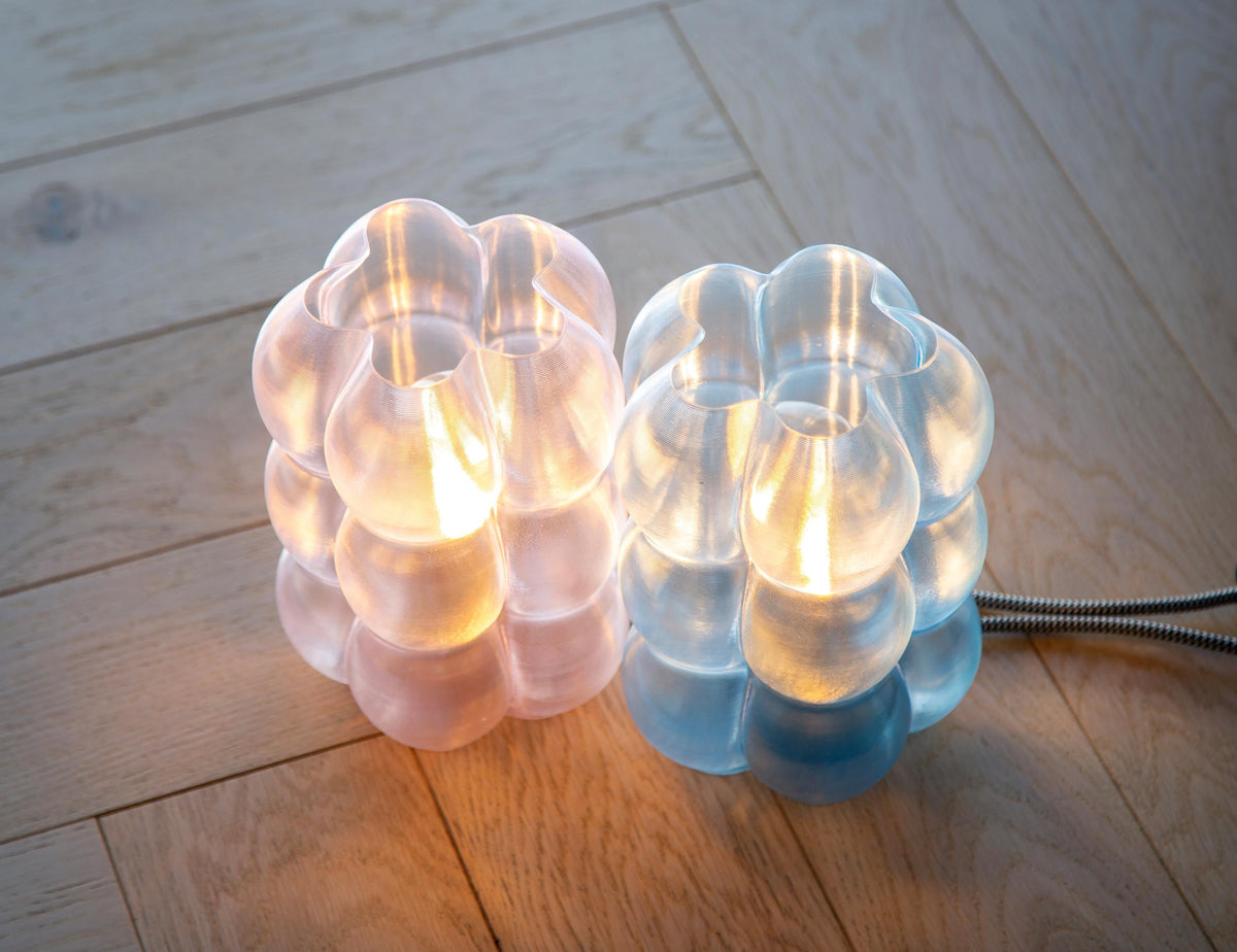 Two modern bubble cluster table lamps on a light wood floor, one glowing with warm pinkish light and the other with cool blue light, with a metal cord visible on the right.