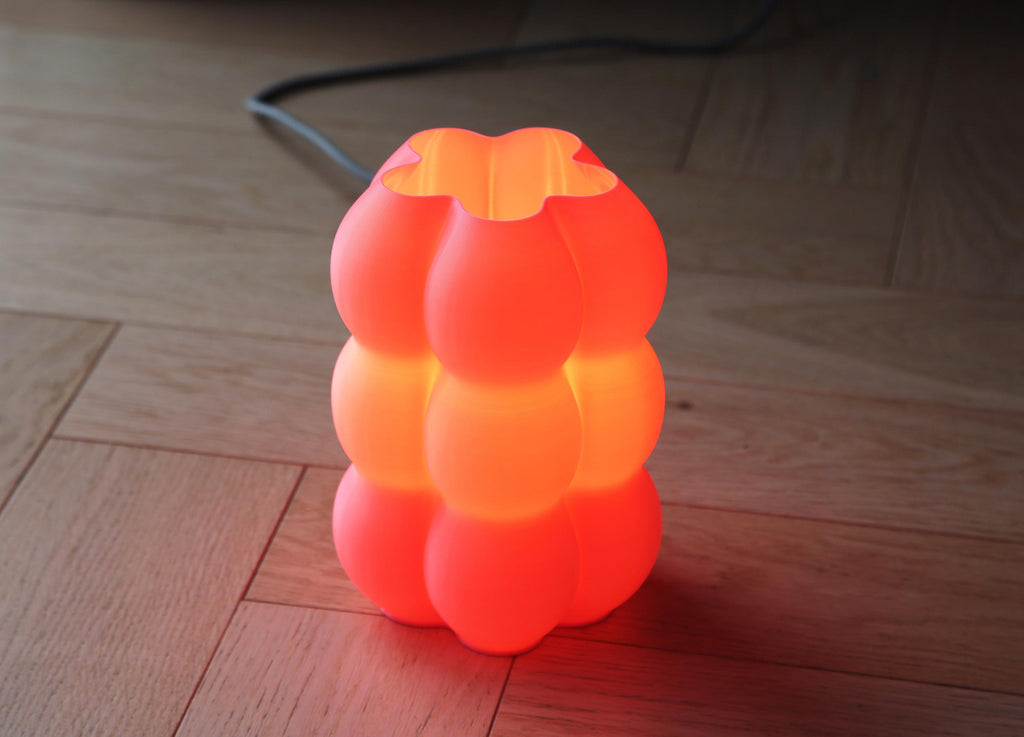 A glowing orange and red bubble-style table lamp with a translucent sculptural shade, placed on a wooden floor with its power cord visible in the background.