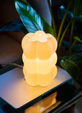 A glowing, sculptural table lamp with a warm yellow light, featuring a stacked, bubble-like organic design, shown on a reflective surface with green plants in the background.