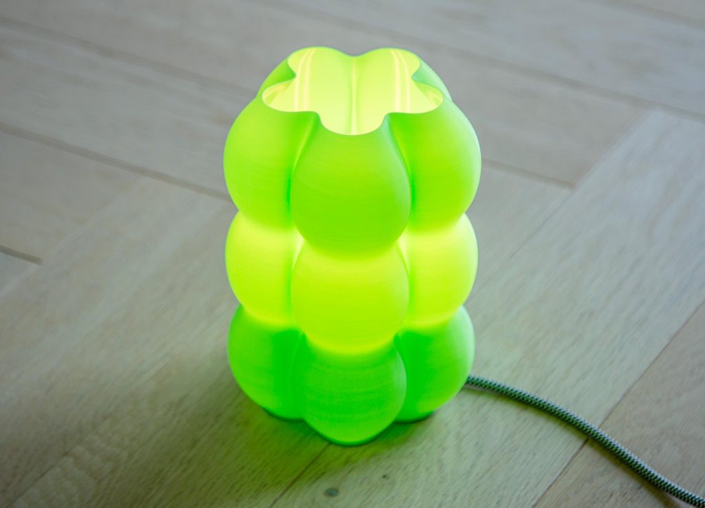 A bright neon green, bulb-shaped table lamp with a glowing interior, set on a light wood floor with its power cord visible on the right.