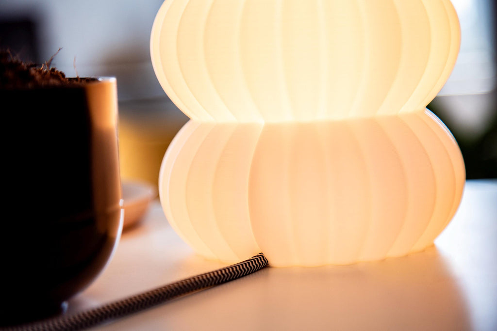 A close-up of a glowing two-tiered table lamp with a ribbed, pleated design, casting a warm yellow light next to a dark planter on a white surface.